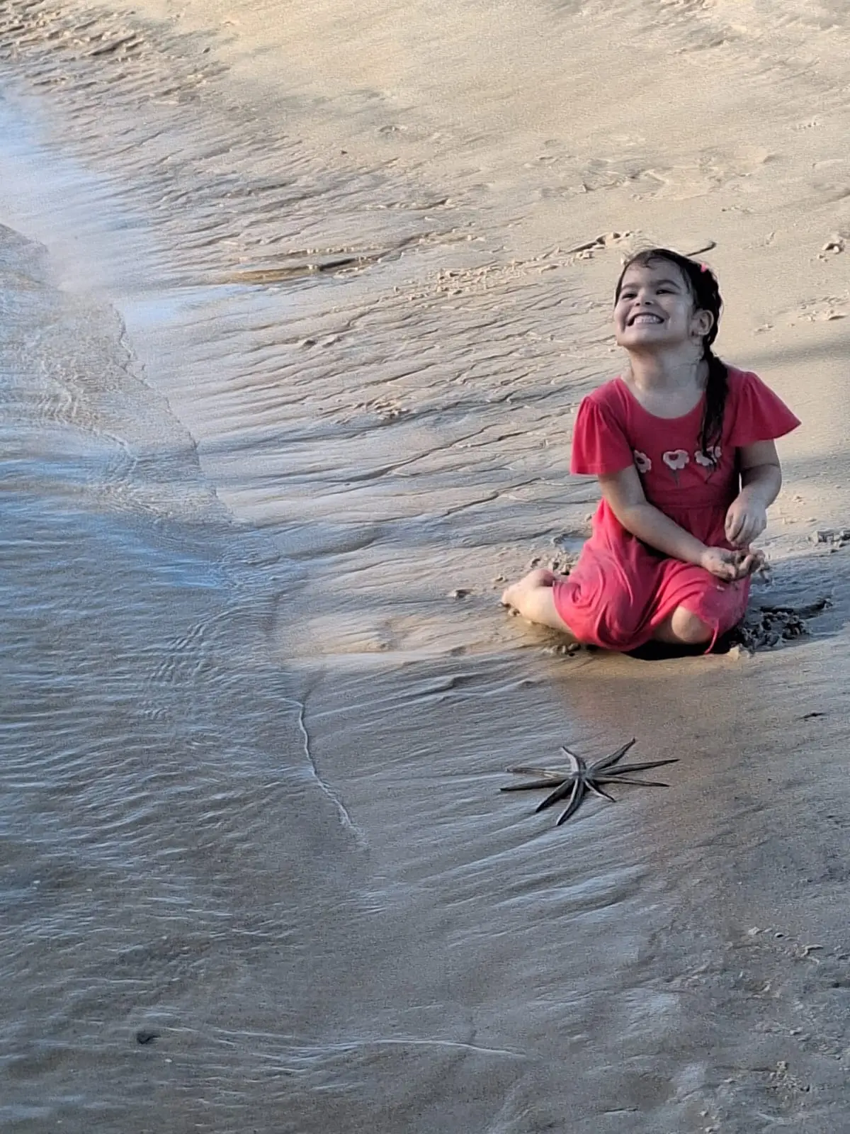 Criança sentada na beira da água, rindo, numa praia de areia clara